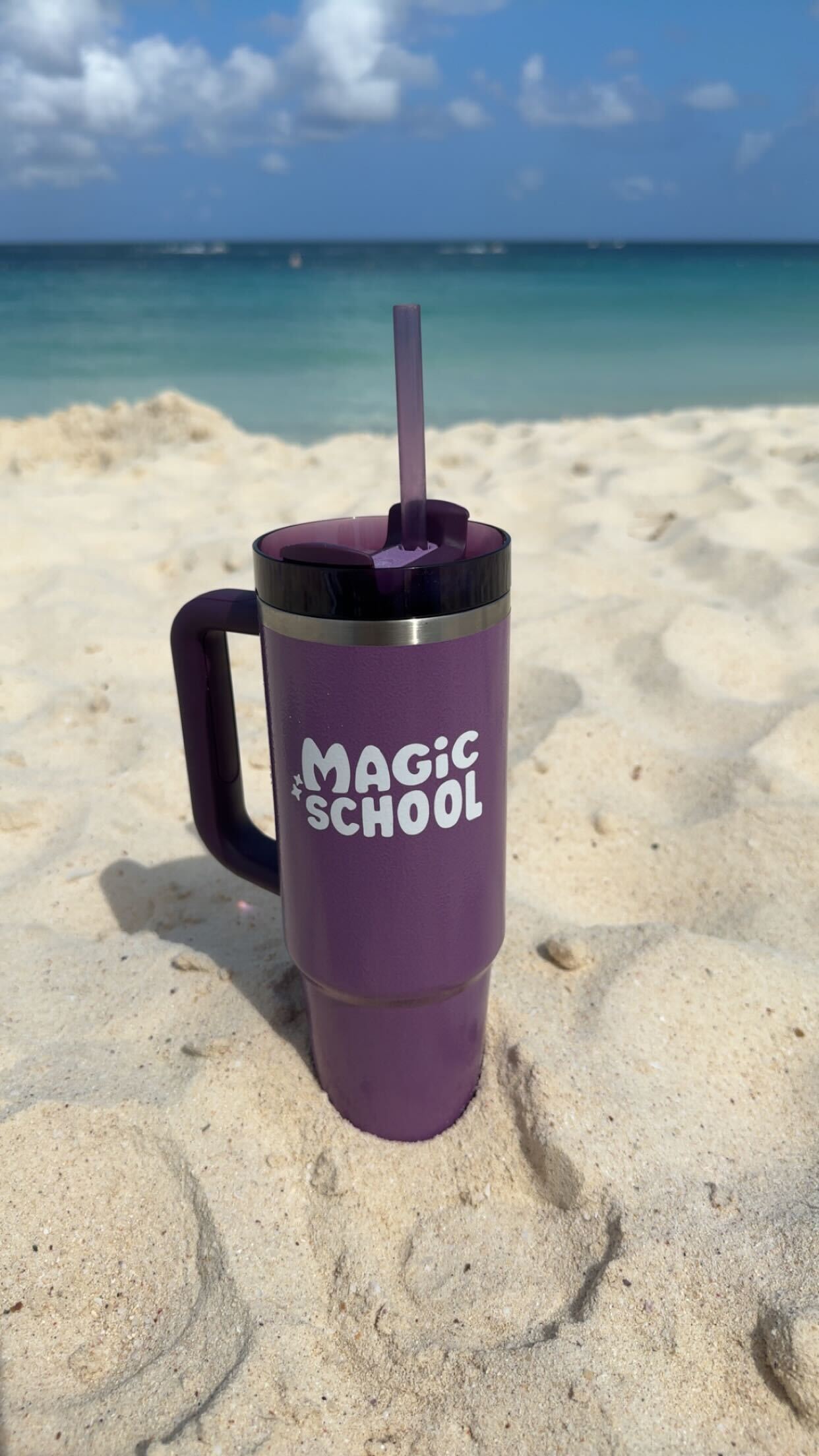 Purple MagicSchool branded Stanley tumbler in the sand on the beach